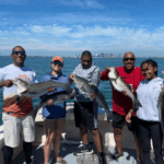 Five fishermen hold up large striped bass on a boat, with a scenic waterfront city skyline and clear blue sky in the background.