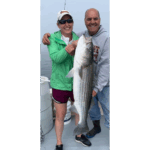 A person wearing a green jacket holds a large striped bass on a fishing boat, with calm water and fishing gear in the background.